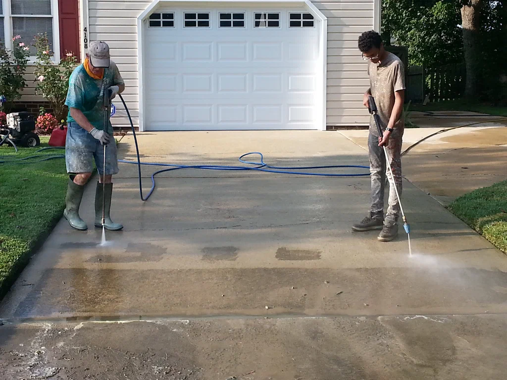 Two people pressure washing a concrete driveway in front of a white garage.