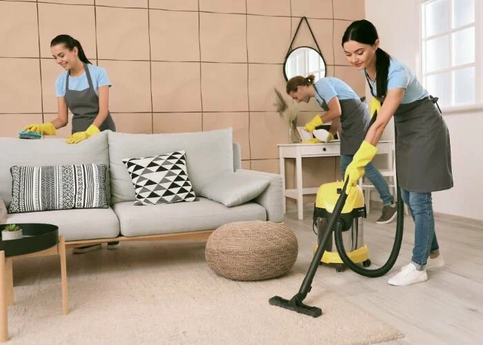 Three professional cleaners in grey aprons vacuuming the carpet and dusting furniture in a living room.