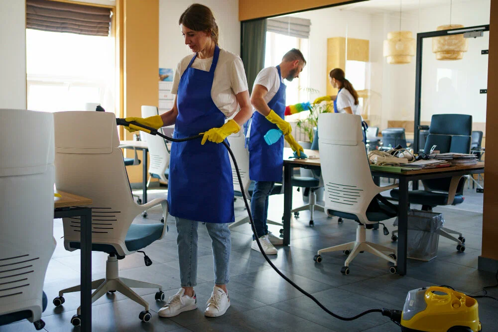 A cleaner in a blue apron vacuums a white office chair while colleagues clean desks in a modern office.