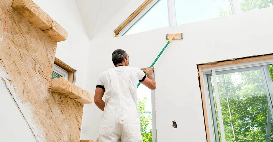 A worker in white overalls sanding a white wall with a long-handled tool in a room under construction.