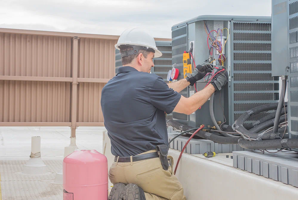 HVAC technician in a hard hat and gloves repairing an industrial AC unit on a rooftop.