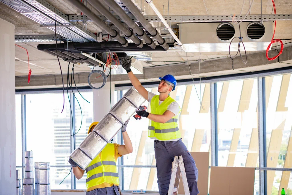 Two construction workers in safety gear installing insulated piping in an unfinished ceiling.