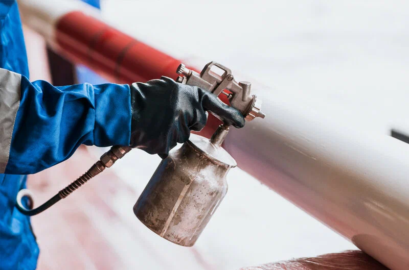 A person in a blue uniform and black gloves uses a spray gun to paint a red pipe white.