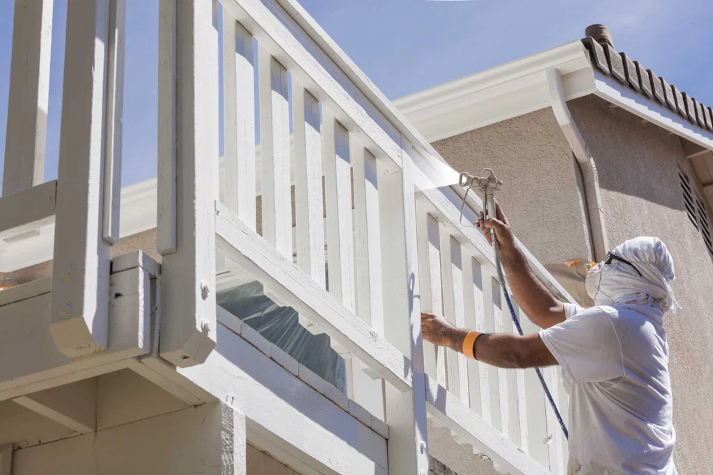 Painter using a spray gun to apply white paint to a wooden deck railing.