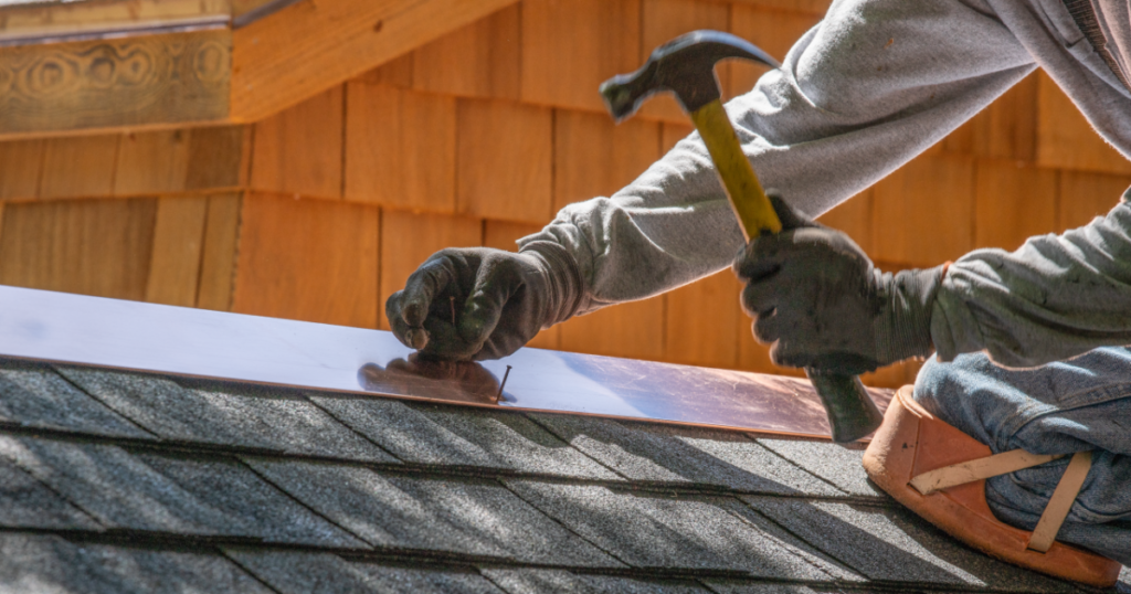 A worker in gloves uses a hammer to nail metal flashing onto a shingled roof.