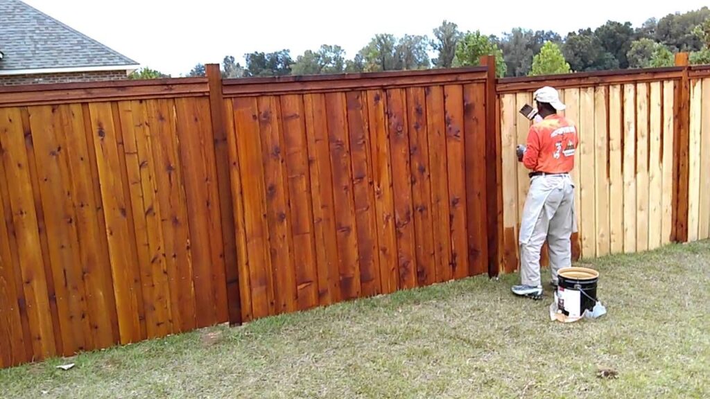 A worker in an orange shirt staining a wooden fence with brown stain using a brush.