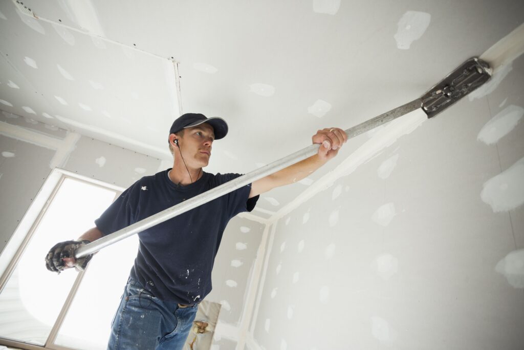 A man in a navy t-shirt and cap uses a long-handled sander to smooth a ceiling patch.