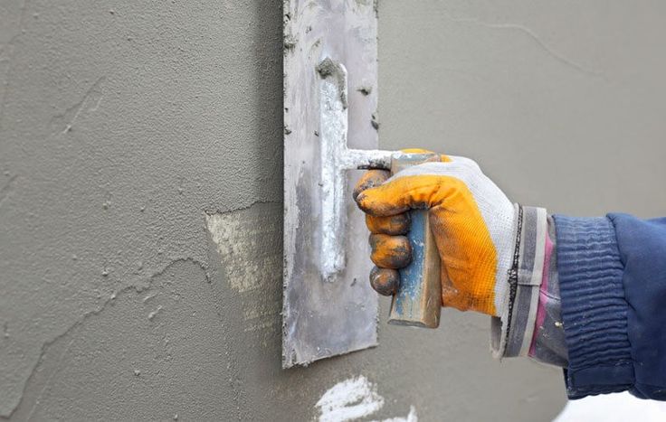 A gloved hand uses a metal trowel to smooth grey plaster on a wall.