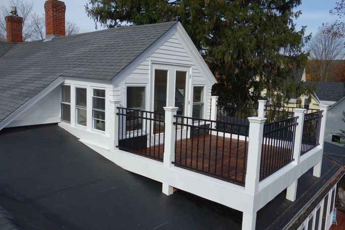 Rooftop deck with dark wood flooring, white posts, and black railings attached to a white dormer on a house.