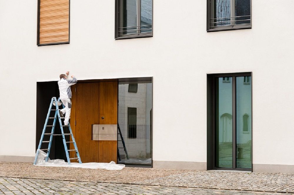 Painter on a blue ladder working on the facade of a white building above a wooden door.