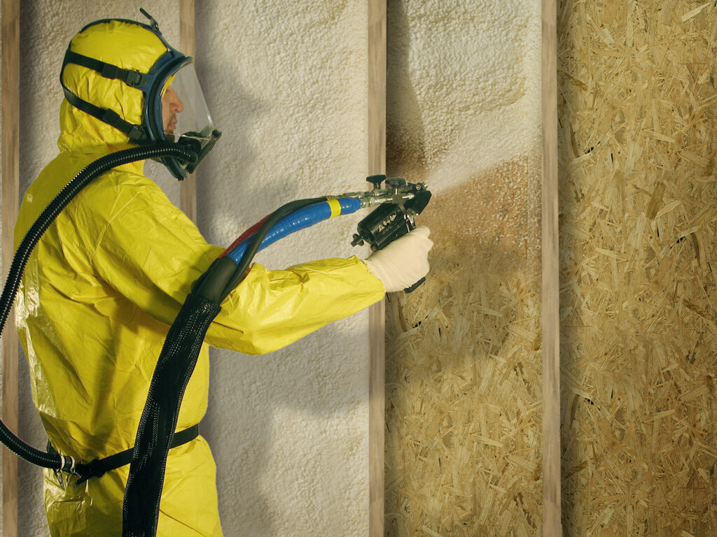 Worker in a yellow hazmat suit and respirator spraying spray foam insulation into a wall cavity.
