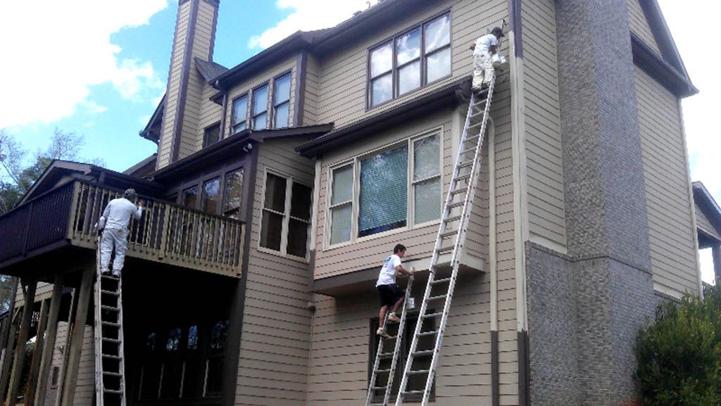 Three painters using tall ladders to paint the exterior siding and balcony railings of a large, multi-story house.