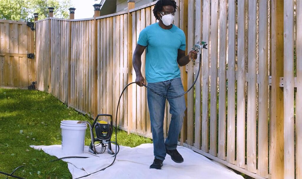 A man wearing a mask and goggles uses a paint sprayer on a wooden backyard fence.