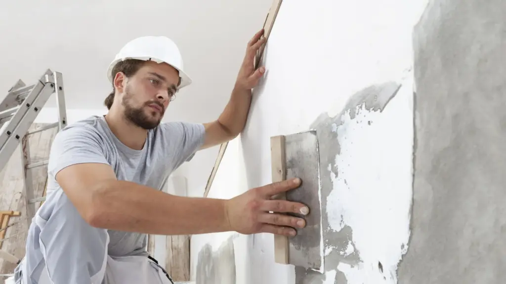 Construction worker in a hard hat smoothing plaster on a wall with a trowel.