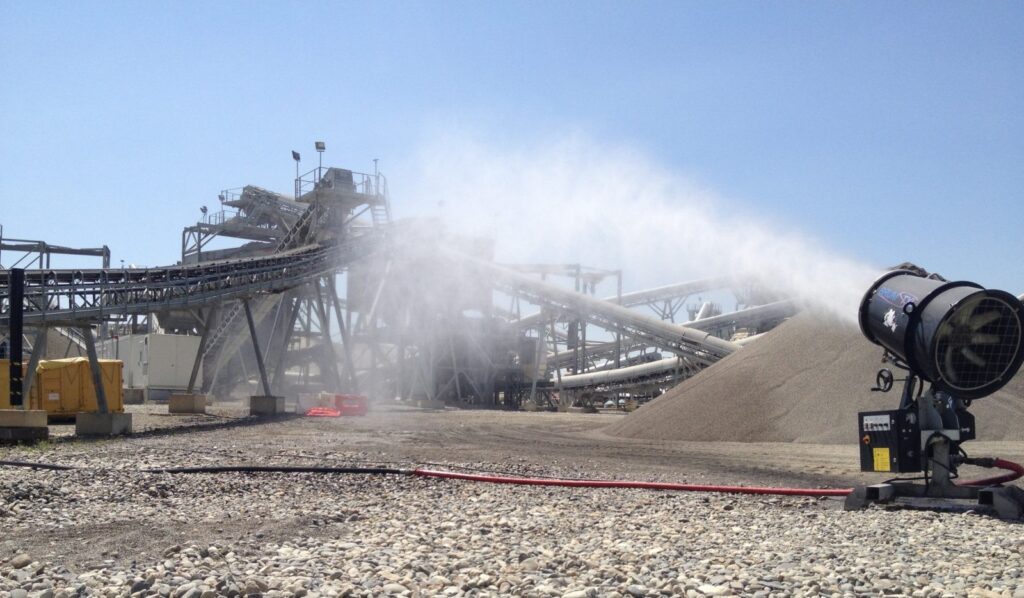 Dust suppression cannon spraying mist at a quarry site with conveyor belts and gravel piles.