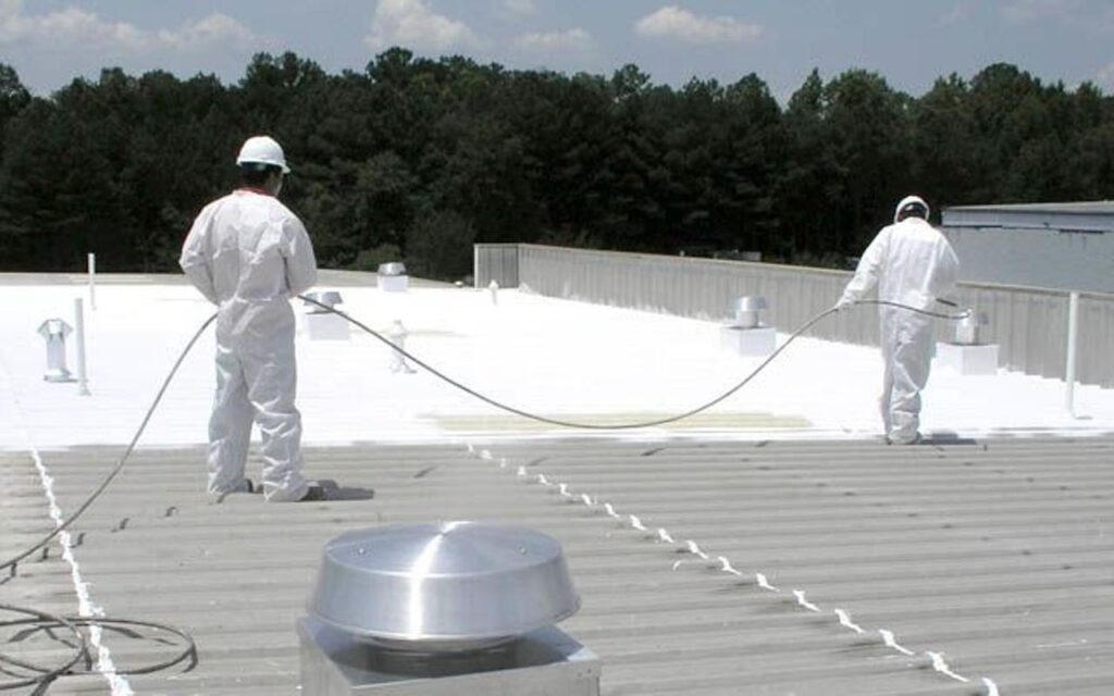 Two workers in white protective suits apply a white sealant to a corrugated metal roof.