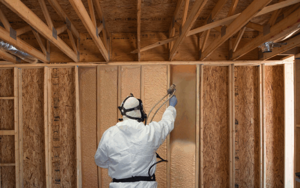 Worker in white protective suit spraying foam insulation into wooden wall studs of a house under construction.