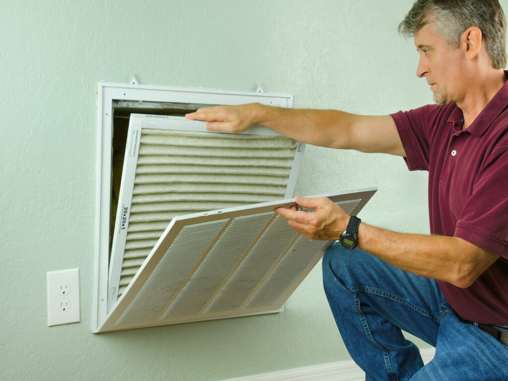 Man sliding a new pleated air filter into a wall-mounted vent.