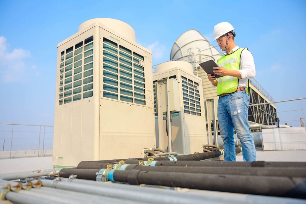 Technician in safety gear using a tablet to monitor industrial HVAC units on a rooftop.