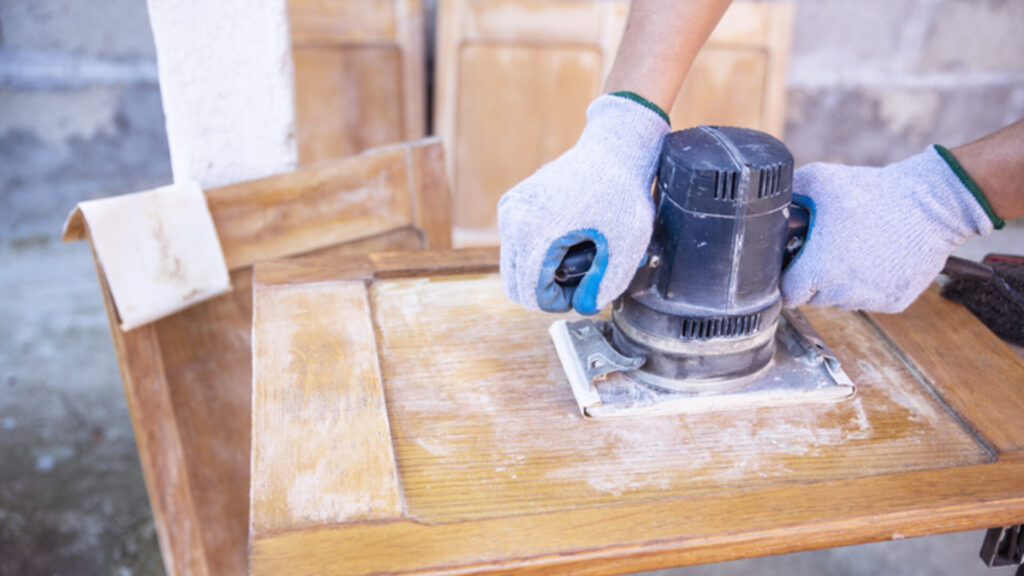 Gloved hands using a power sander on a wooden furniture panel.