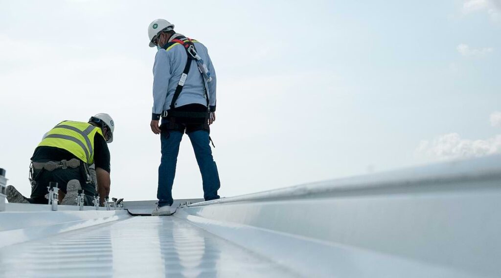 Two construction workers in safety gear and hard hats installing a metal roof.