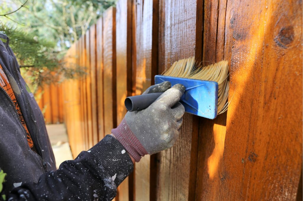 Gloved hand applying brown stain to a wooden fence with a blue brush.