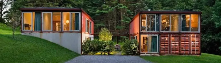 Two modern red shipping container homes with large windows, connected by a walkway in a lush forest setting.