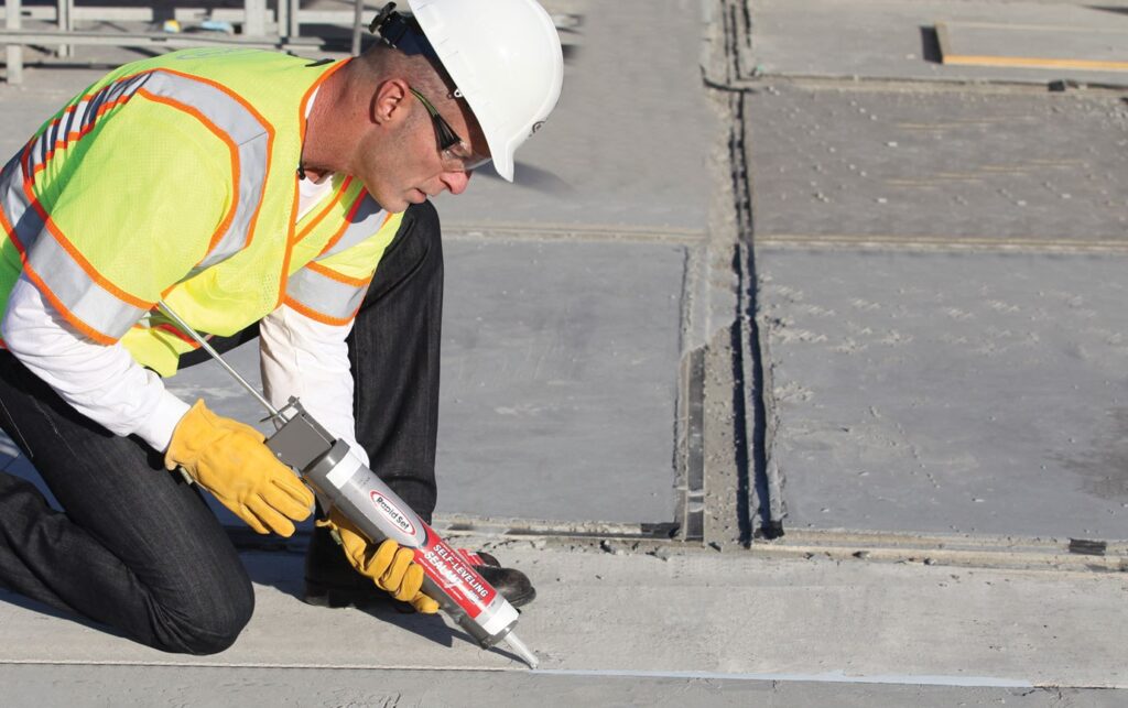 A worker in safety gear uses a caulk gun to apply self-leveling sealant to a concrete joint.