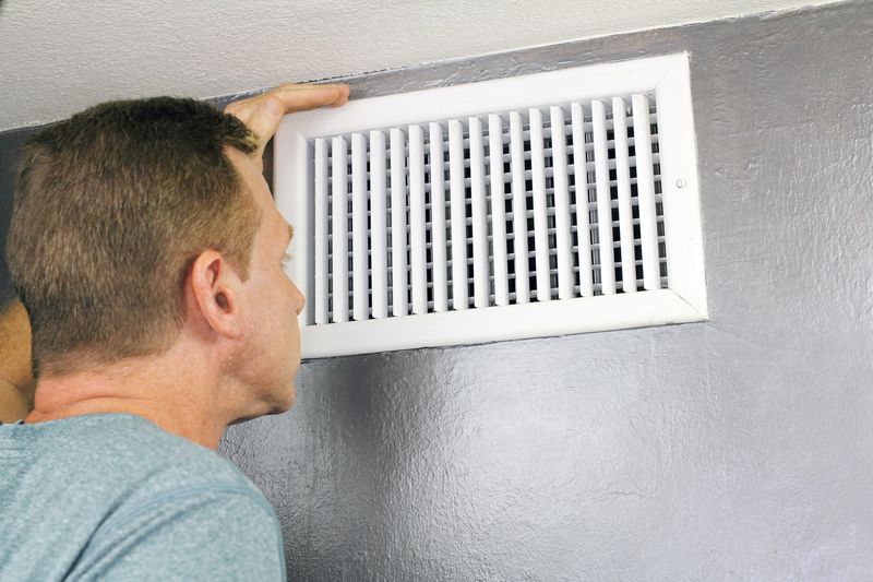 A man inspects a white air vent mounted on a gray wall.