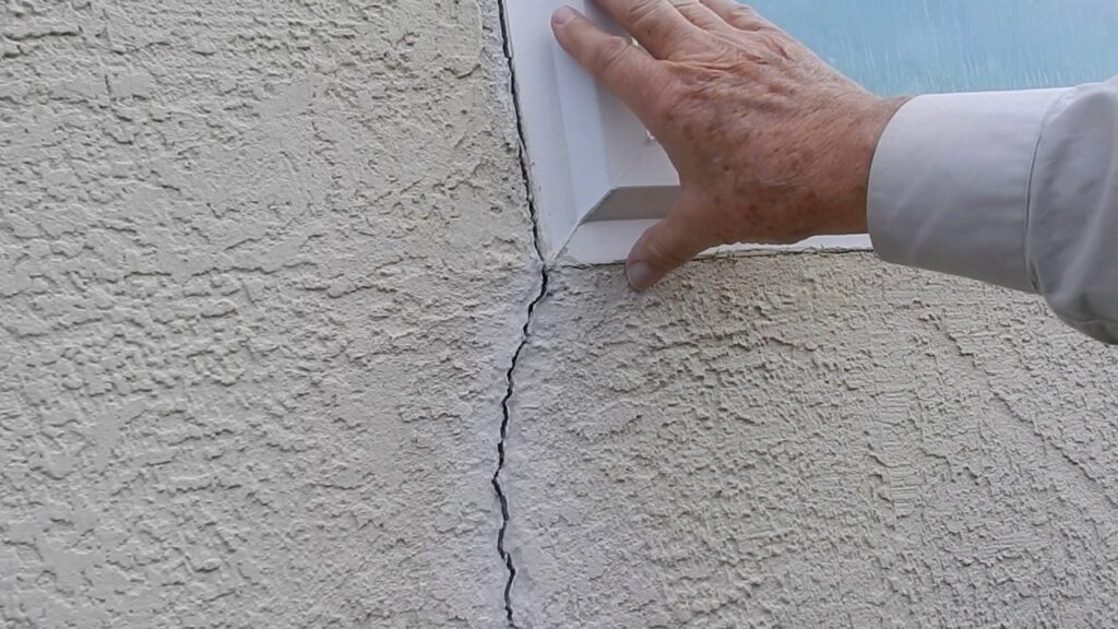 A hand points to a vertical crack in a stucco exterior wall next to a white window frame.