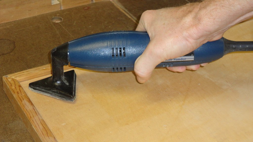 A hand holding a blue detail sander to sand the inside corner of a wooden panel.