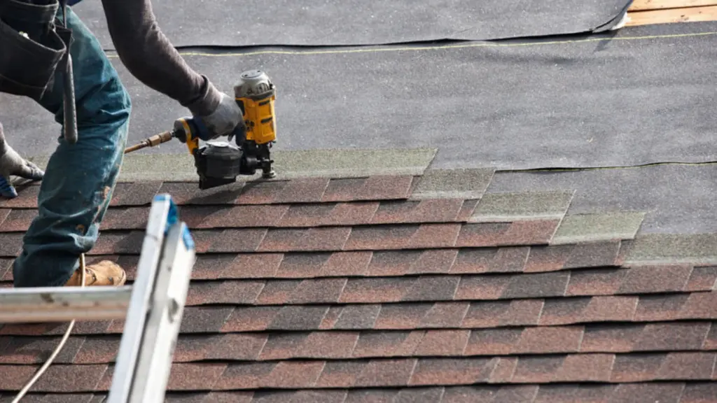 Roofer installing brown asphalt shingles using a yellow pneumatic nail gun.
