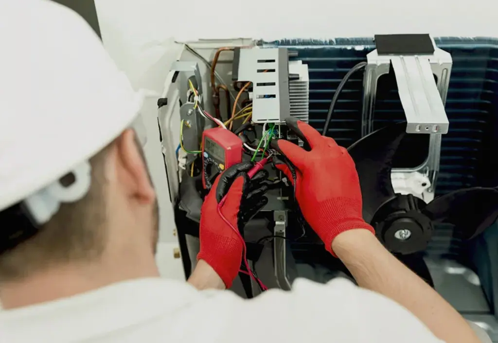 Technician in red gloves using a multimeter to test the internal wiring of an open air conditioning unit.