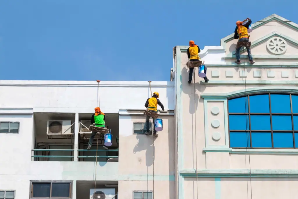 Four painters suspended by ropes paint the exterior walls of a building against a clear blue sky.