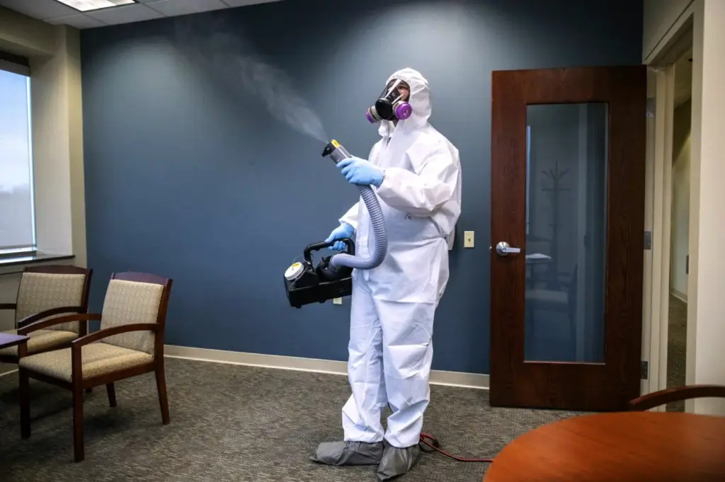Person in a white hazmat suit and respirator using a fogging machine to disinfect an office.