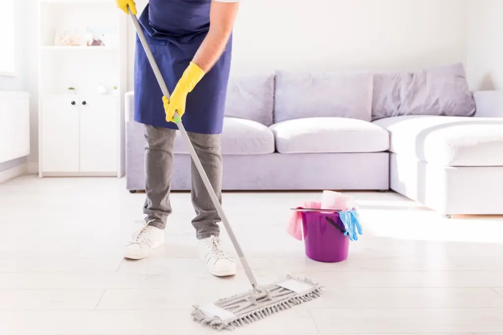 Person in a blue apron and yellow gloves mopping a living room floor next to a purple cleaning bucket.