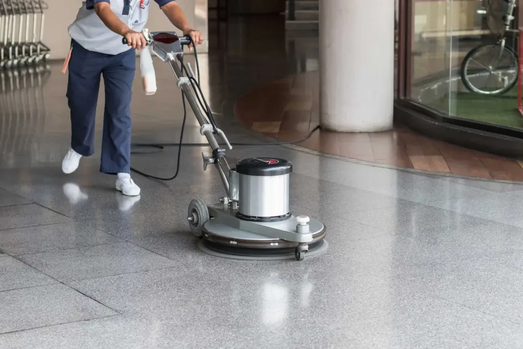 A janitor uses a floor buffer machine to polish a shiny stone floor in an indoor hallway.