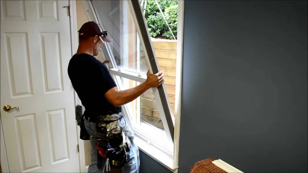A man in a tool belt installs a white window frame into a house wall.