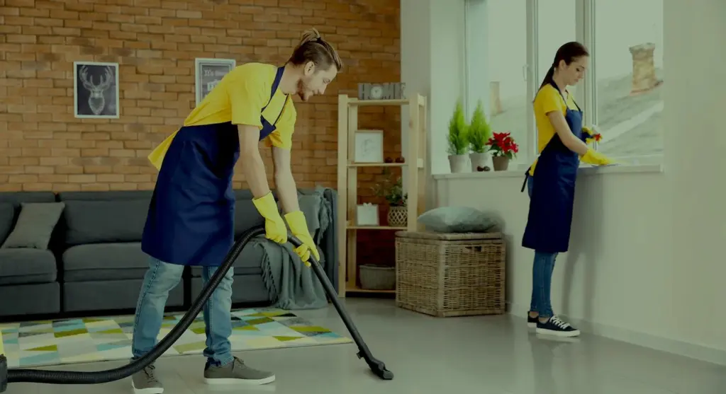 Two professional cleaners in yellow shirts and blue aprons; one vacuums the floor while the other cleans a window sill.