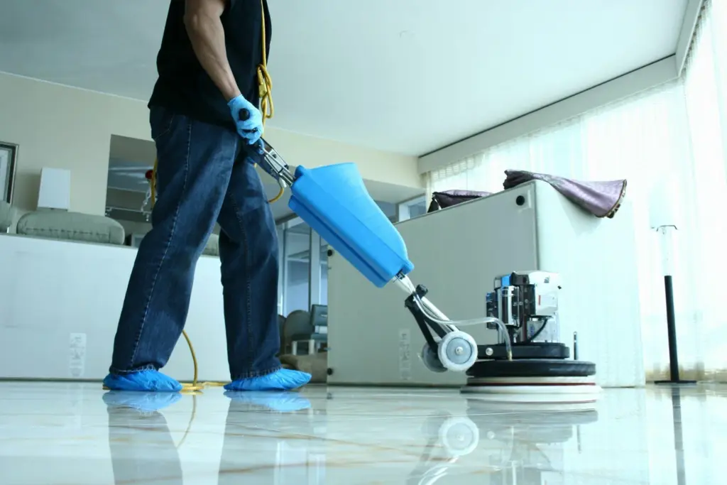 A person wearing blue gloves and shoe covers uses a floor cleaning machine on a polished tile floor.