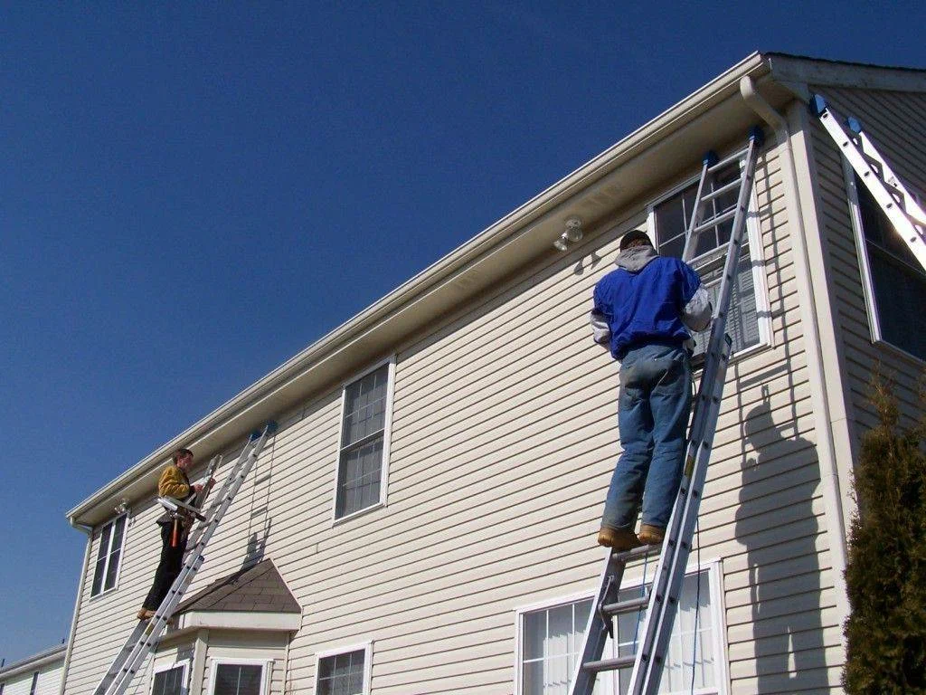 Two workers on ladders working on the exterior siding of a two-story house.