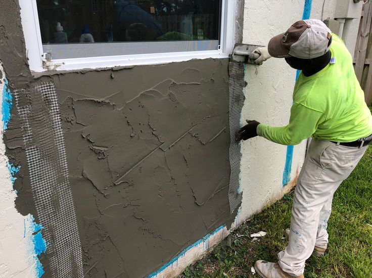 Construction worker applying grey stucco to an exterior wall beneath a window.