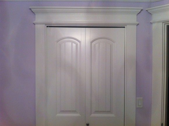 White double closet doors with arched panels and ornate white trim against a purple wall.