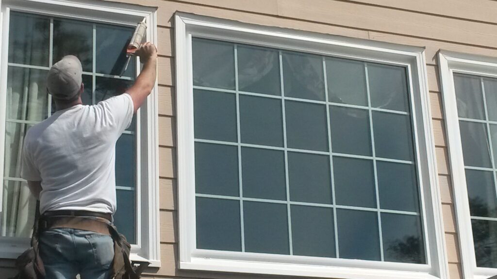 Worker using a caulk gun to seal the edge of a white window frame on a tan house.