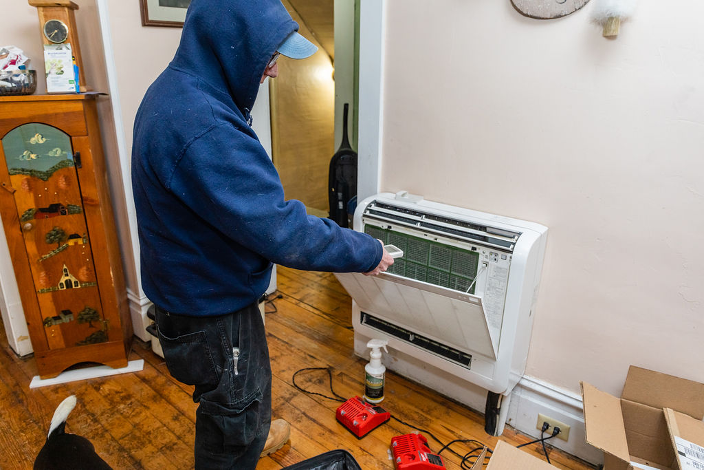 A person in a blue hoodie servicing a wall-mounted HVAC unit with the front panel open.