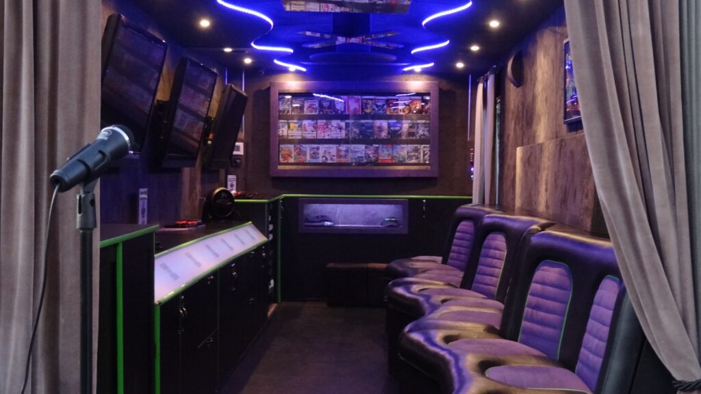 Interior of a mobile gaming trailer with purple neon ceiling lights, a wall of video games, and purple seating.