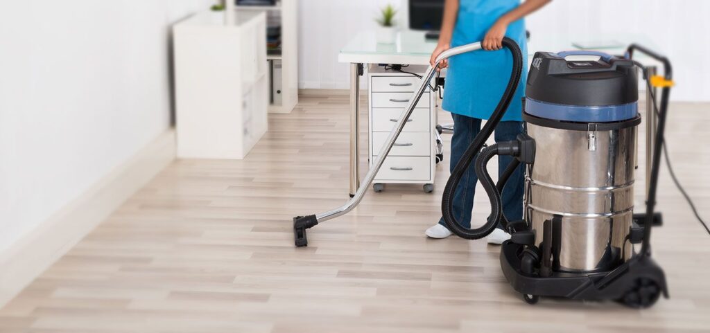 A professional cleaner using an industrial vacuum on a light wood floor in an office.