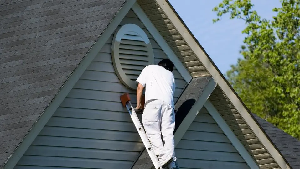 A painter on a ladder works on a circular attic vent on a house's exterior siding.