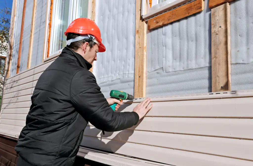 Construction worker in a red hard hat using a drill to install beige vinyl siding on a building exterior.