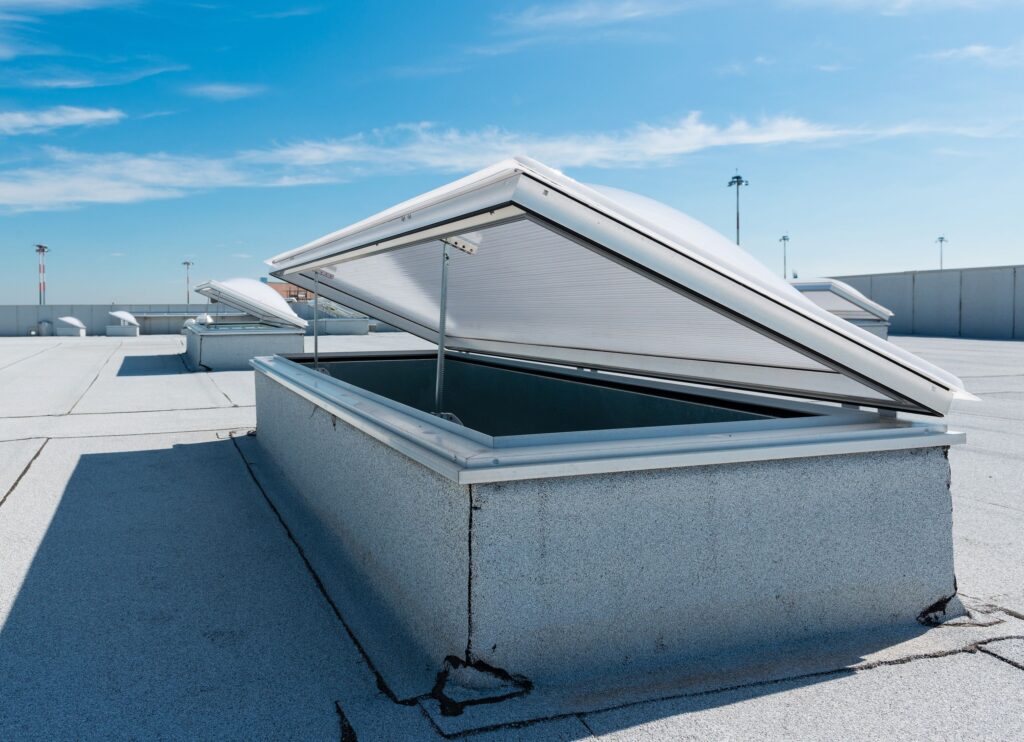 Open white roof hatch on a gray flat roof under a clear blue sky.